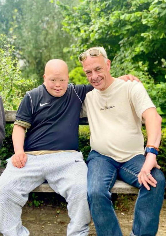 Twee mannen zitten naast elkaar op een bank in een groene omgeving, met een vriendelijke uitstraling en een ontspannen houding. De man links draagt een zwart sportshirt en grijze trainingsbroek, terwijl de man rechts een lichte T-shirt en jeans aan heeft.