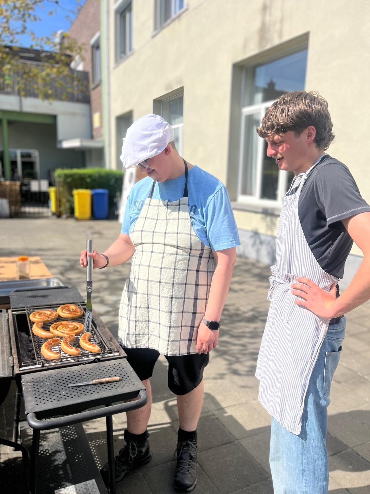 Twee mensen in schort staan buiten bij een grill, waarbij één persoon een gerecht omdraait met een spatel.