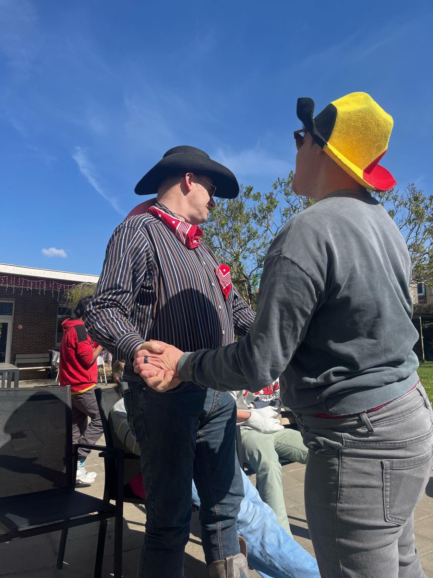 Twee mensen staan elkaar hand in hand, met een blauwe lucht boven hen. De man rechts draagt een zwart en geel hoed, terwijl de man links een zwart hoed en een gestreepte shirt met een rode bandana heeft. Achter hen zijn andere mensen en bomen zichtbaar.