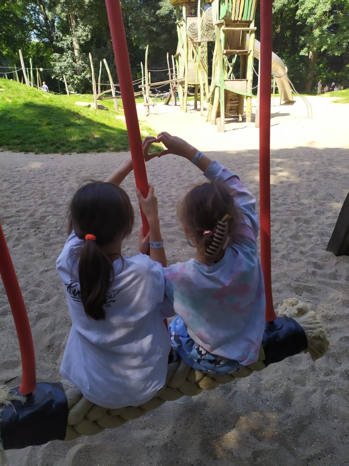Twee kinderen zitten op een schommel in een speeltuin en vormen met hun handen een hartvorm. Achter hen zijn speeltuinstructuren zichtbaar te zien, zoals een glijbaan en klimplekken.