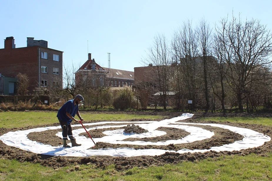 Een persoon aan het werk in een open veld, bezig met het graven van een spiraalvormige structuur met een schep, omringd door gebouwen en bomen onder een blauwe lucht.