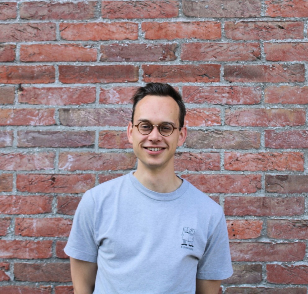 Portret van een man met een grijs T-shirt, bril en een glimlach, staande voor een bakstenen muur.