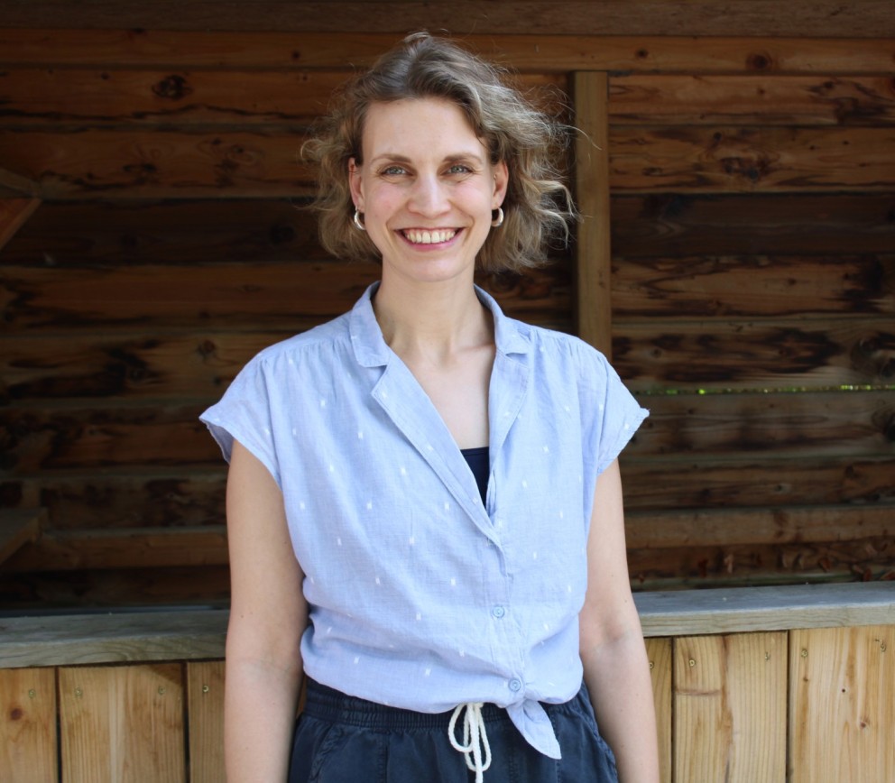 Een vrouw met lichtgekleurde, halflange haren en een glimlach staat voor een houten achtergrond. Ze draagt een lichtblauwe blouse met geknoopte onderkant en een zwarte top eronder.