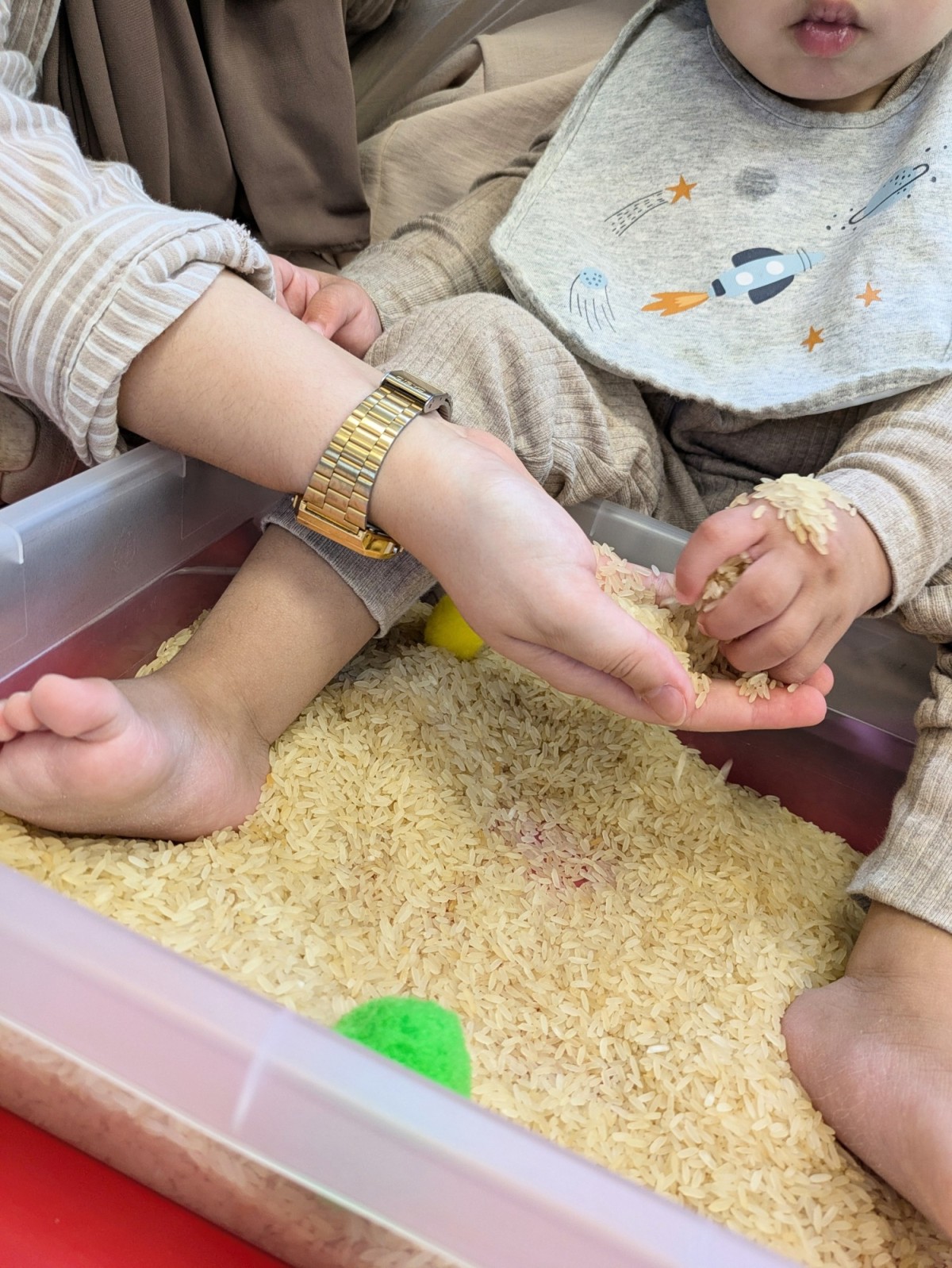 Een kind speelt met rijst in een plastic bak, terwijl een volwassene een hand op de arm van het kind legt; beide zitten op de grond.