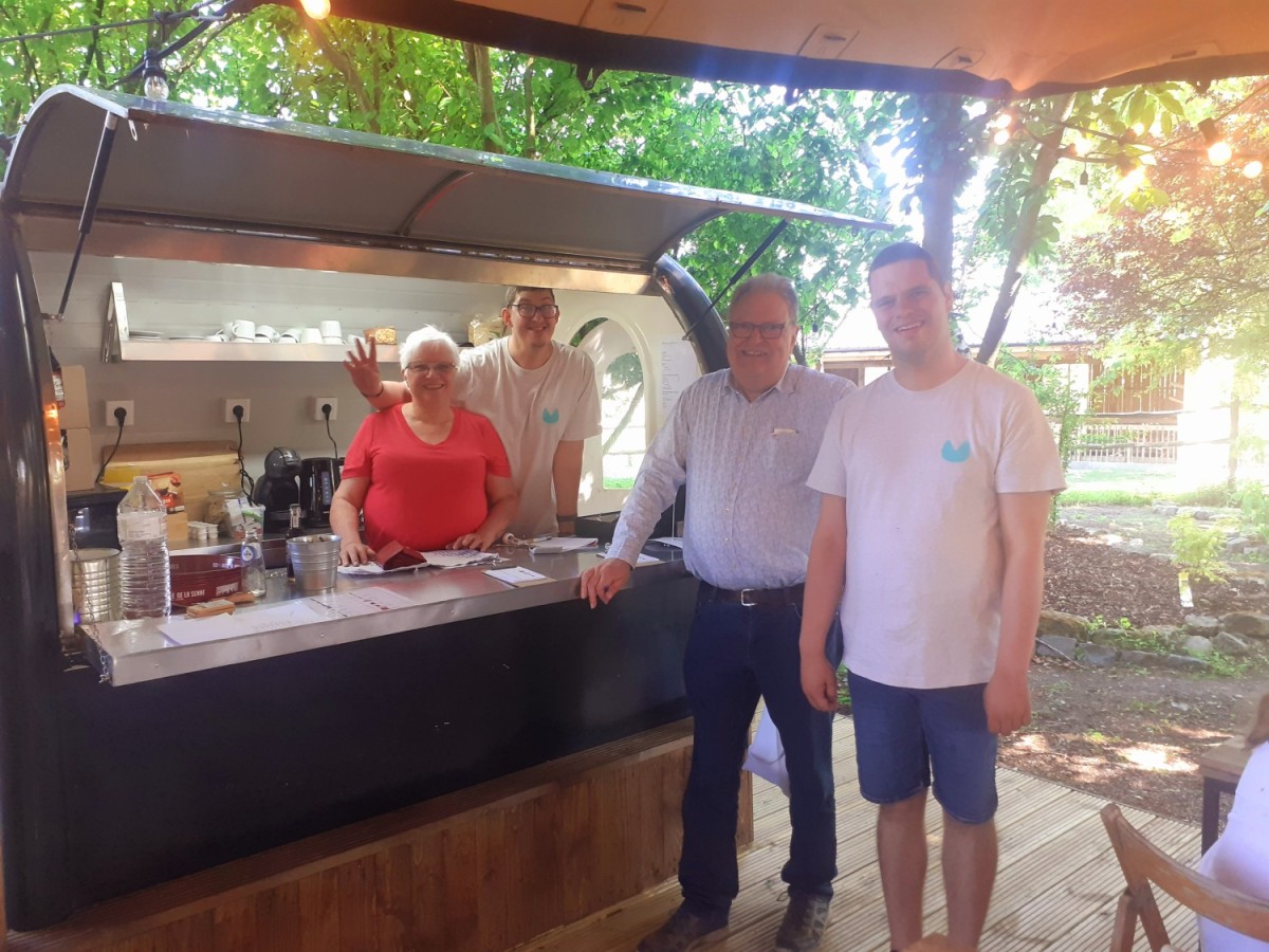 Een groep van vier personen staat achter een eet- en drinkstand. Twee mannen en twee vrouwen lachen en poseren voor de camera, met een groene omgeving op de achtergrond.