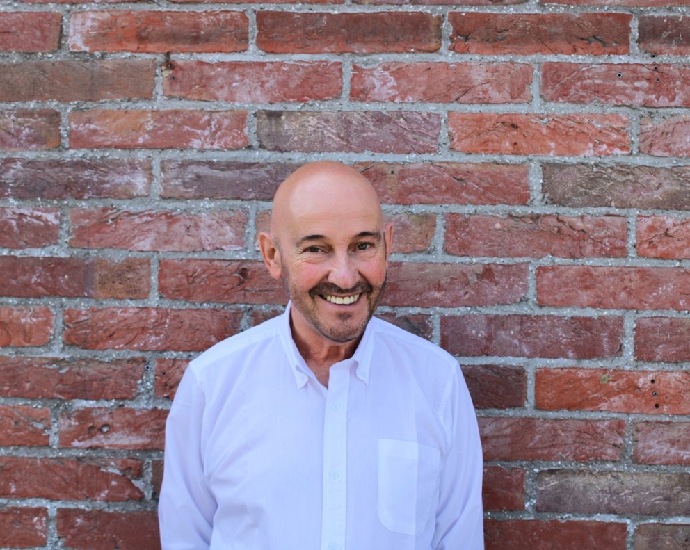 Man met een kaal hoofd en een snor, die glimlacht terwijl hij tegen een bakstenen muur staat. Hij draagt een witte blouse.