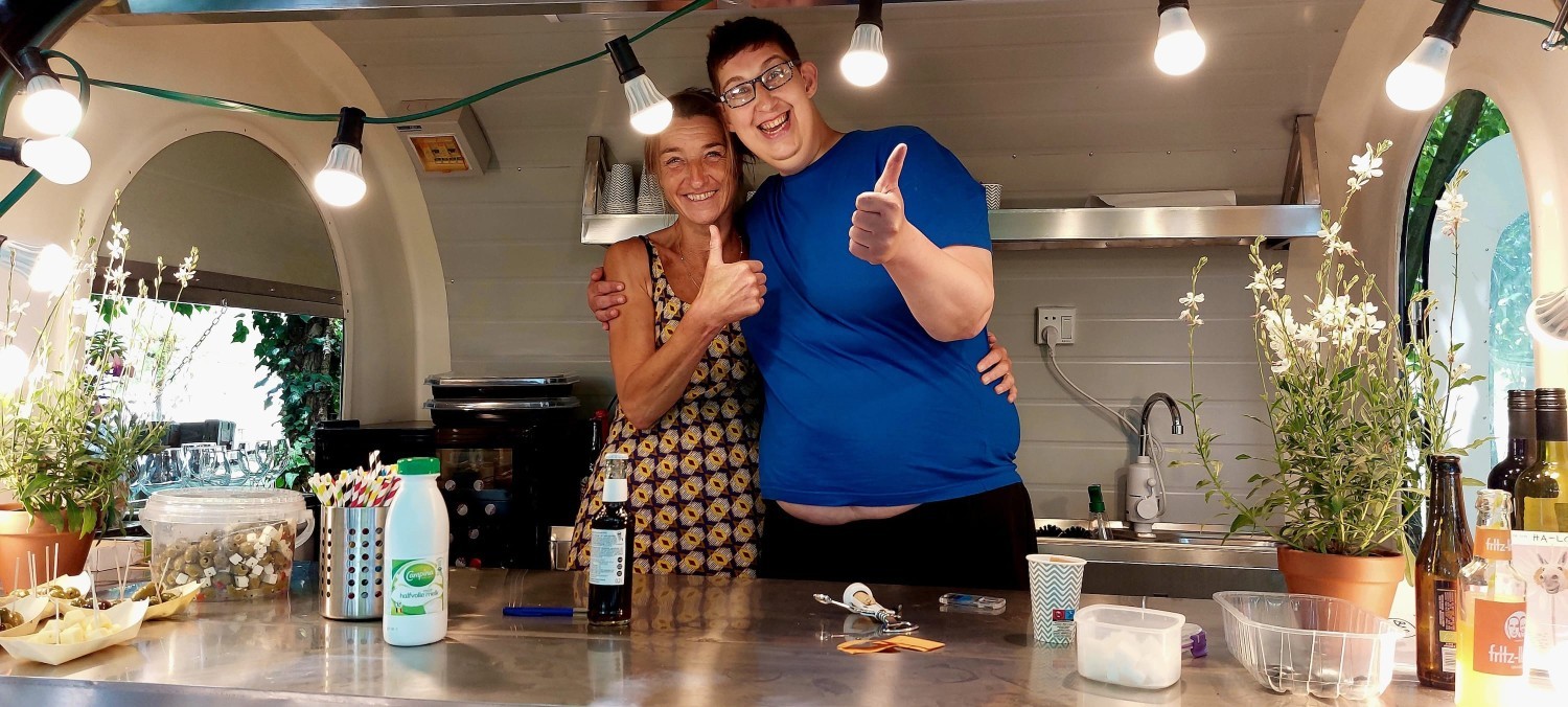 Twee mensen staan in een kleine keuken met een metalen werkblad, omringd door planten en lampen, en geven beide een duim omhoog terwijl ze glimlachen.